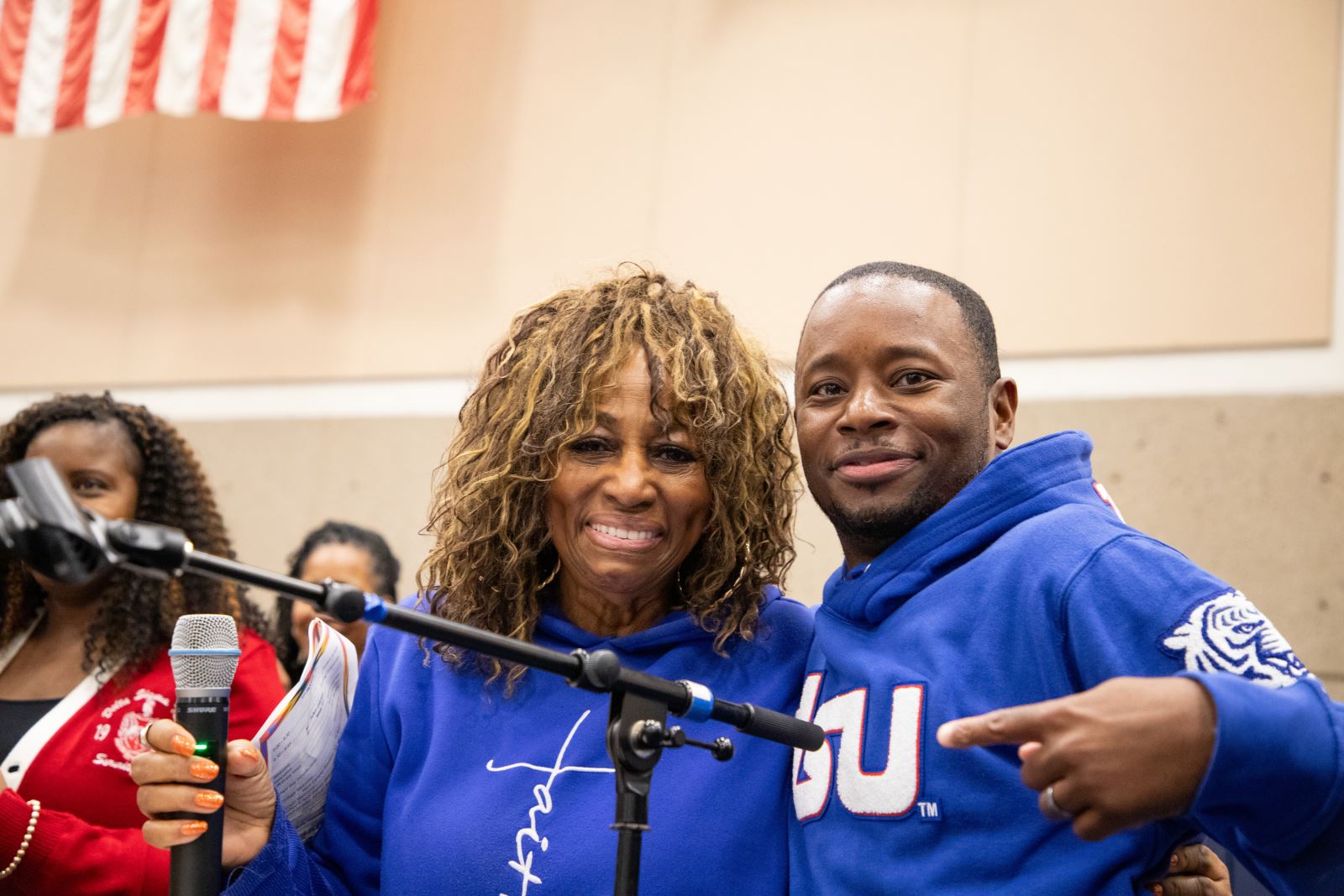 Two speakers, a woman and a man dressed in bright blue hoodies, smile by the microphone.