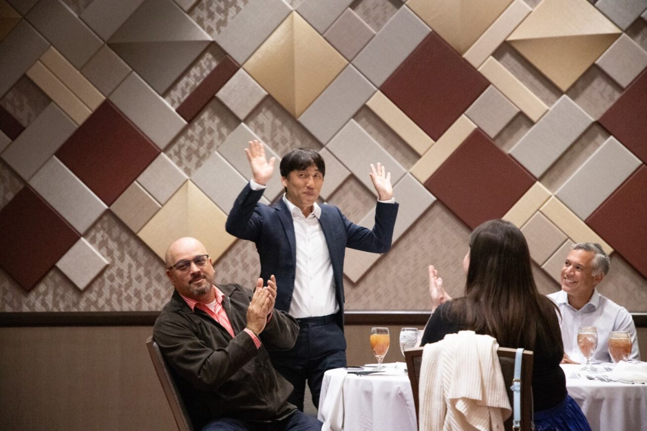 A man raises his hands up in appreciation while a man and a woman clap at the table